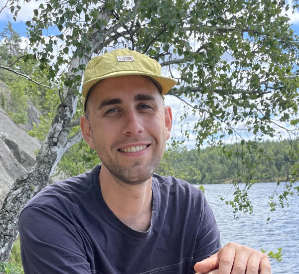 man looking into the camera sitting next to a body of water
